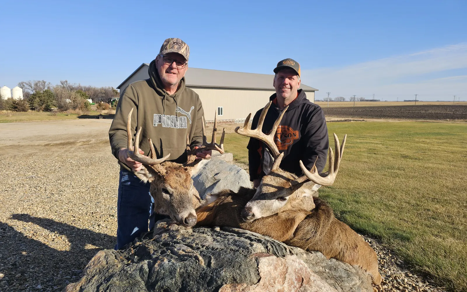 Two hunters kneeling behind two whitetail bucks on a large rock outside a farm shop